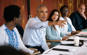 Barack Obama sits at a meeting table and talks vividly surrounded by other people